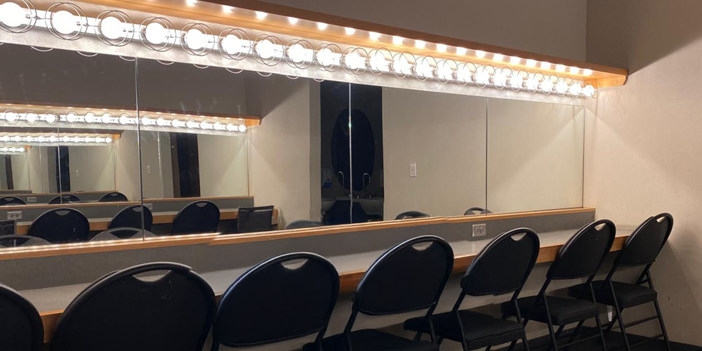 row of bright makeup lights above wall width mirrors adjacent to countertop and dressing room chairs