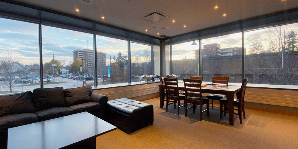 Room with large wrap around windows, dark grey sofa, black ottoman, and a dining table with chairs